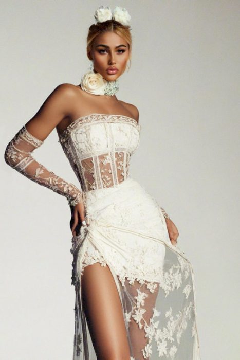 Ein Modeporträt einer Frau in einem weißen, trägerlosen Spitzenkleid mit Korsett-Oberteil. Sie trägt passenden floralen Haarschmuck, ein florales Halsband und lange Spitzenhandschuhe vor einem hellen, neutralen Hintergrund.