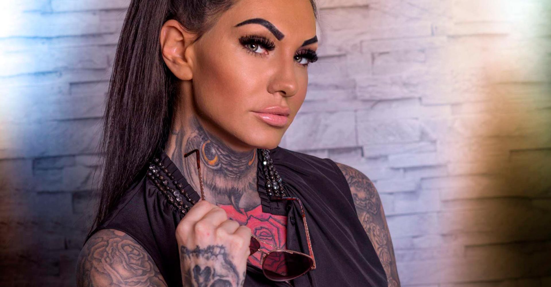 Porträt einer tätowierten Frau mit Sonnenbrille und Pferdeschwanz vor einer steinernen Wand.